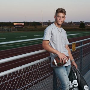 high school senior with letterman jacket and football at prosper practice field