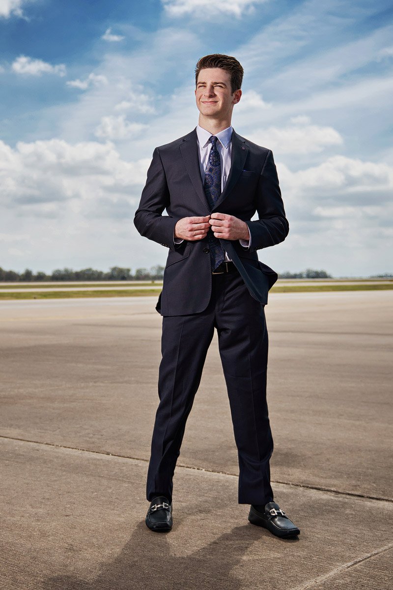 prosper senior photographer photographs student in a suit with blue skies and airport